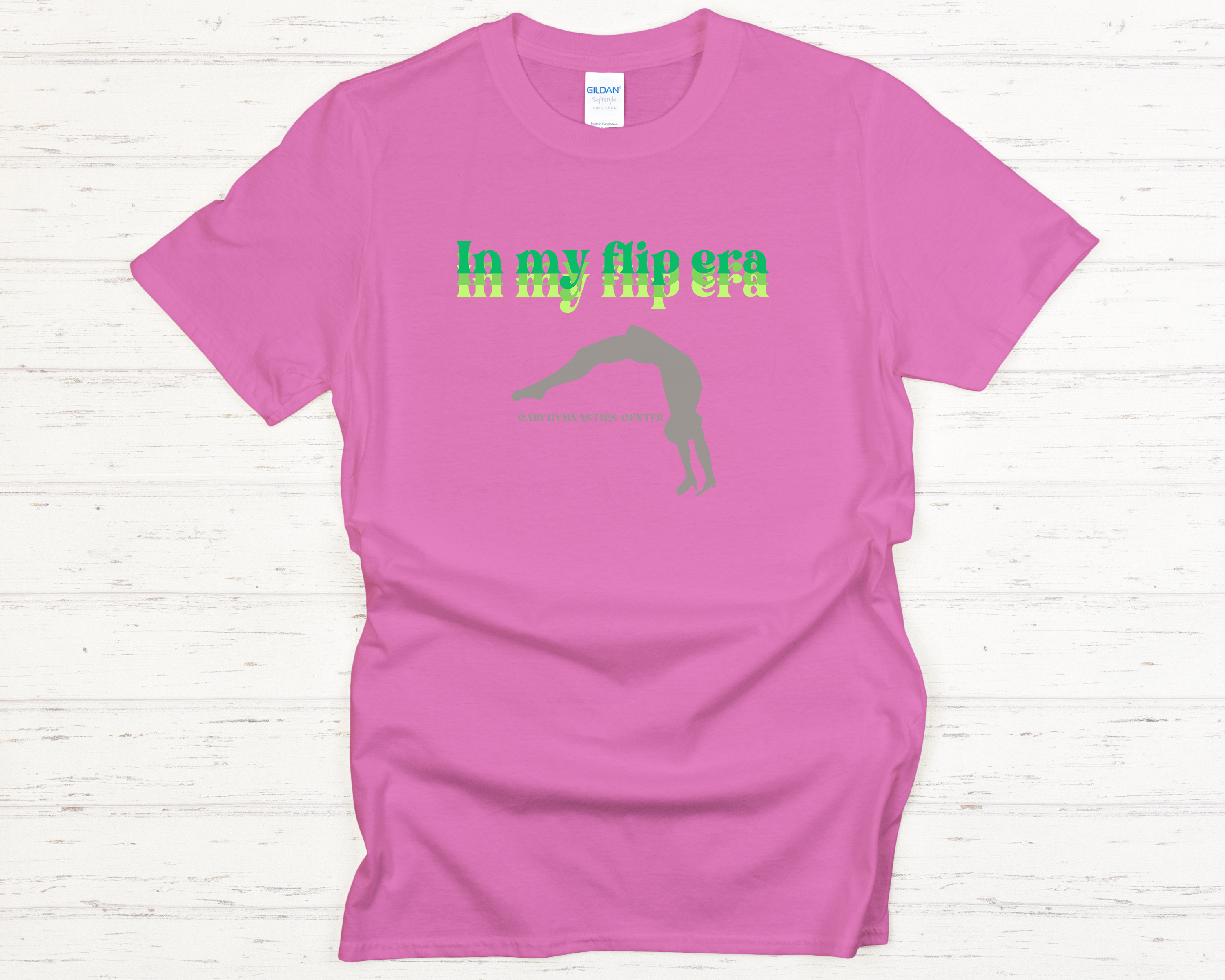 Pink t-shirt with green text and Cary Gymnastics tumbling graphic on a white wooden surface